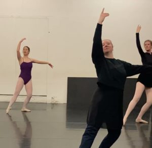 Three ballet dancers in a studio stand in plie with one arm raised