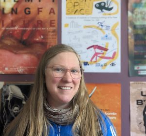 Headshot of a white woman with long blond hair standing in front of a wall of posters